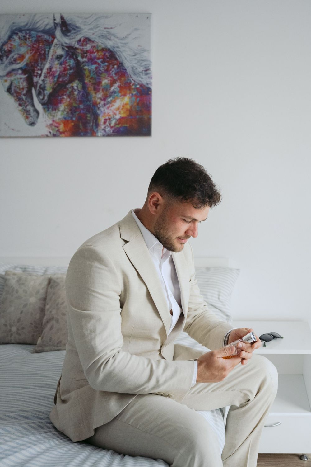 Groom in beige suit checks phone while sitting on gray couch beneath colorful horse artwork.