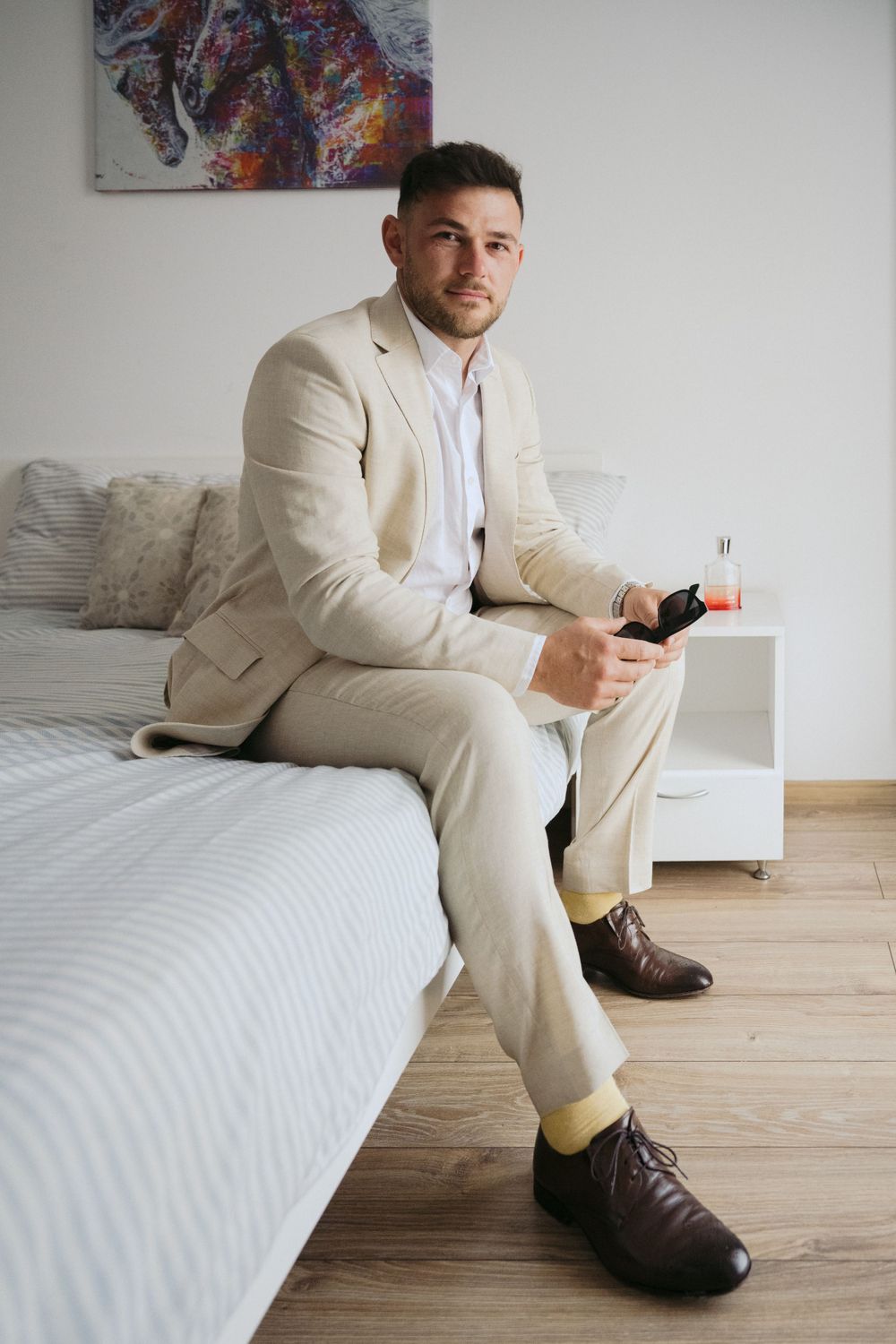 Stylishly dressed person in beige suit and brown shoes sitting on white bed holding phone in modern bedroom.