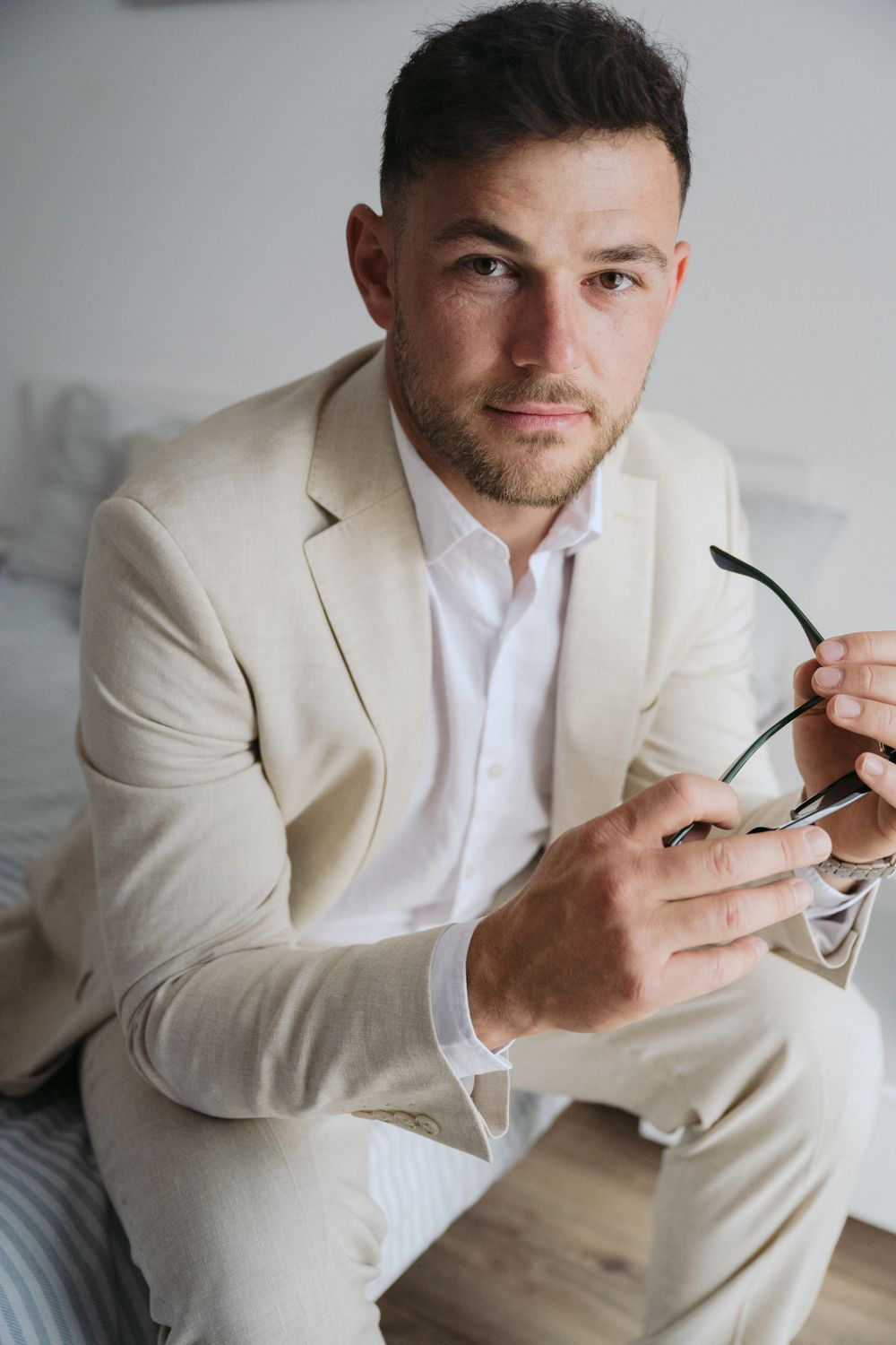 Professional wearing a cream-colored suit and white shirt holding sunglasses while seated indoors.
