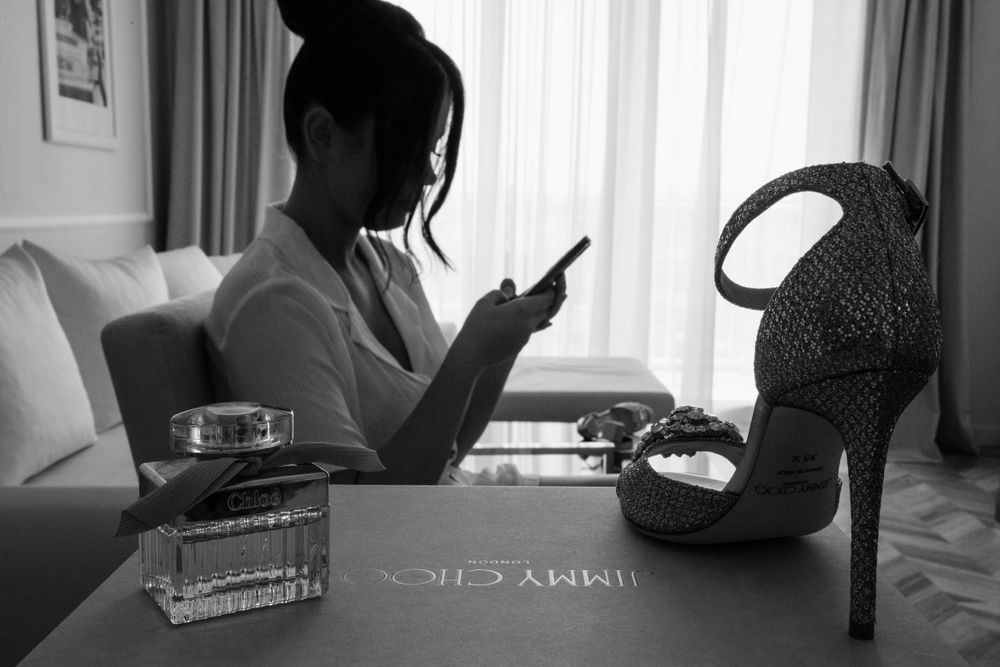 Black and white photo showing silhouette looking at phone next to stiletto heel and perfume bottle on table.