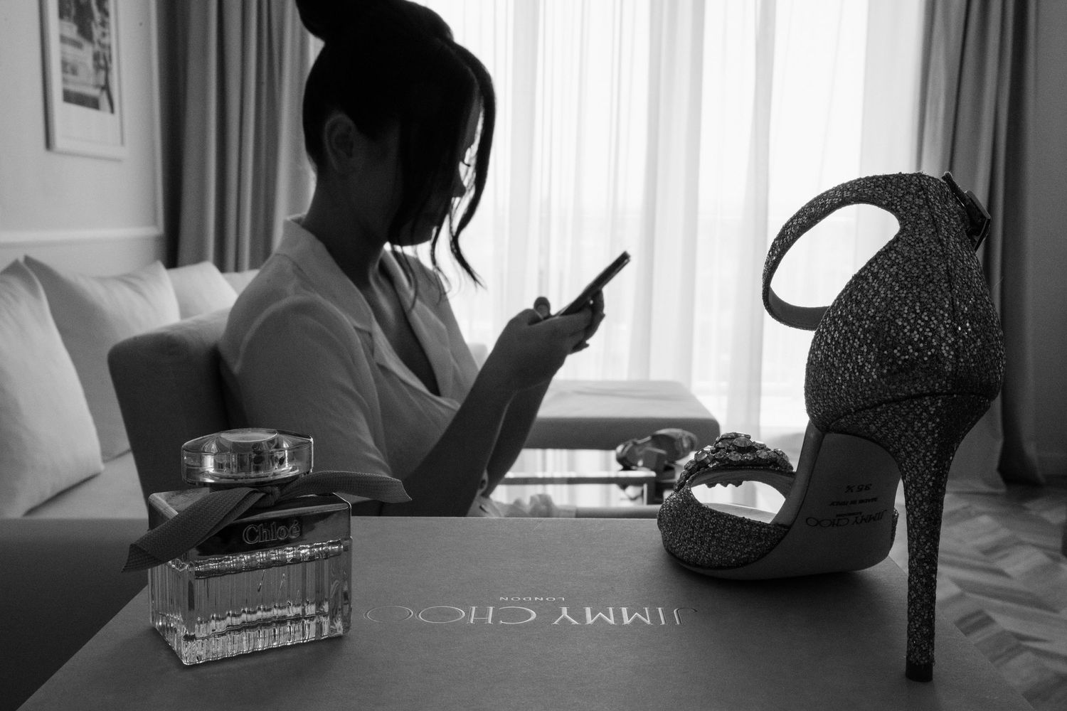 Black and white photo showing silhouette looking at phone next to stiletto heel and perfume bottle on table.