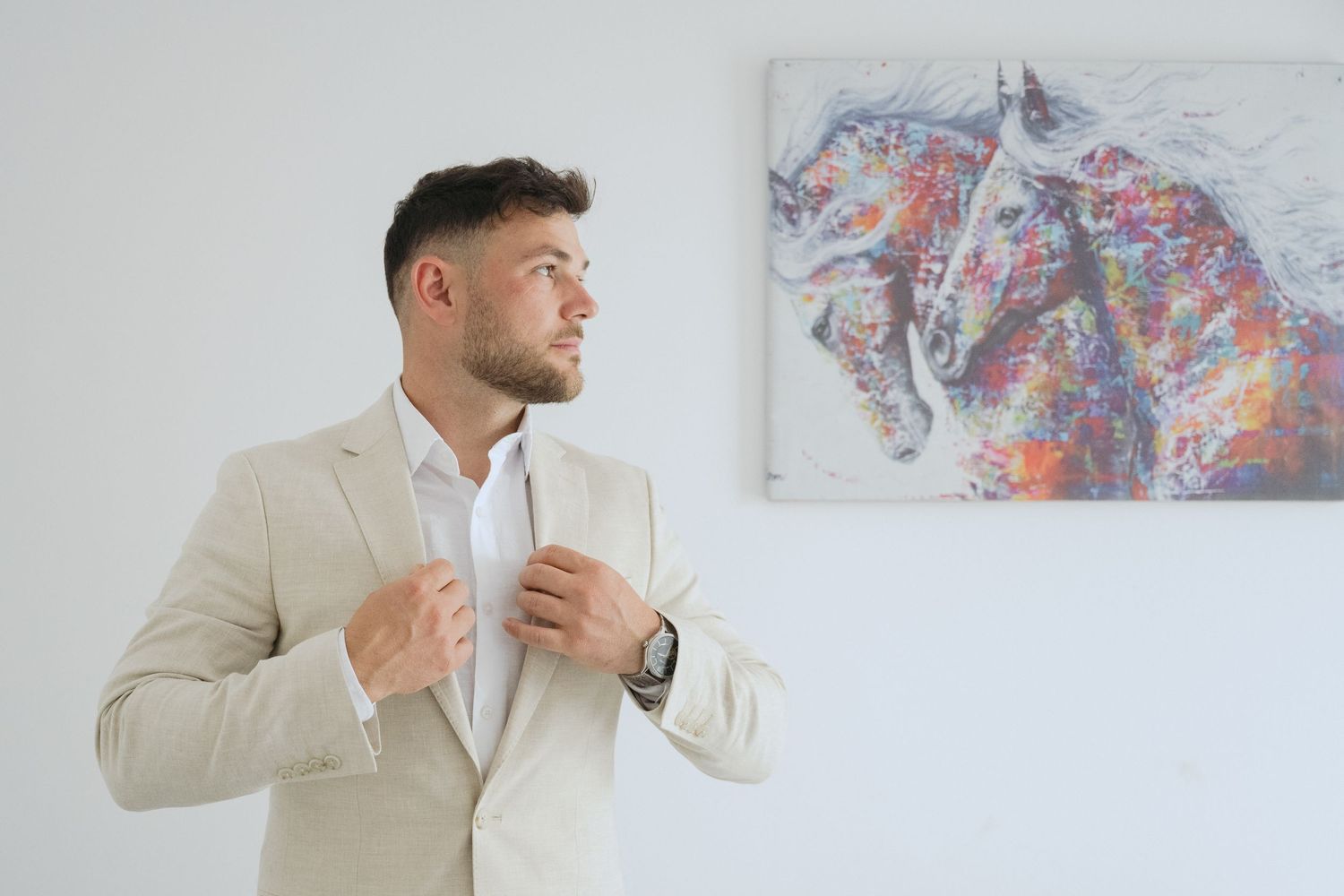 Professional in cream suit jacket stands beside colorful abstract horse painting on white wall.