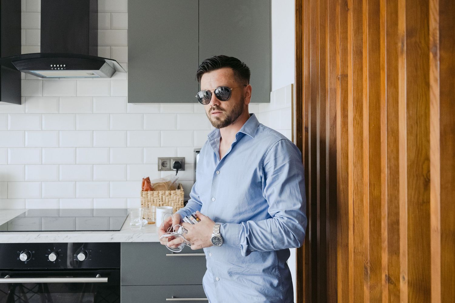 Person in blue button-down shirt and sunglasses pouring a drink in modern kitchen with white tile backsplash.