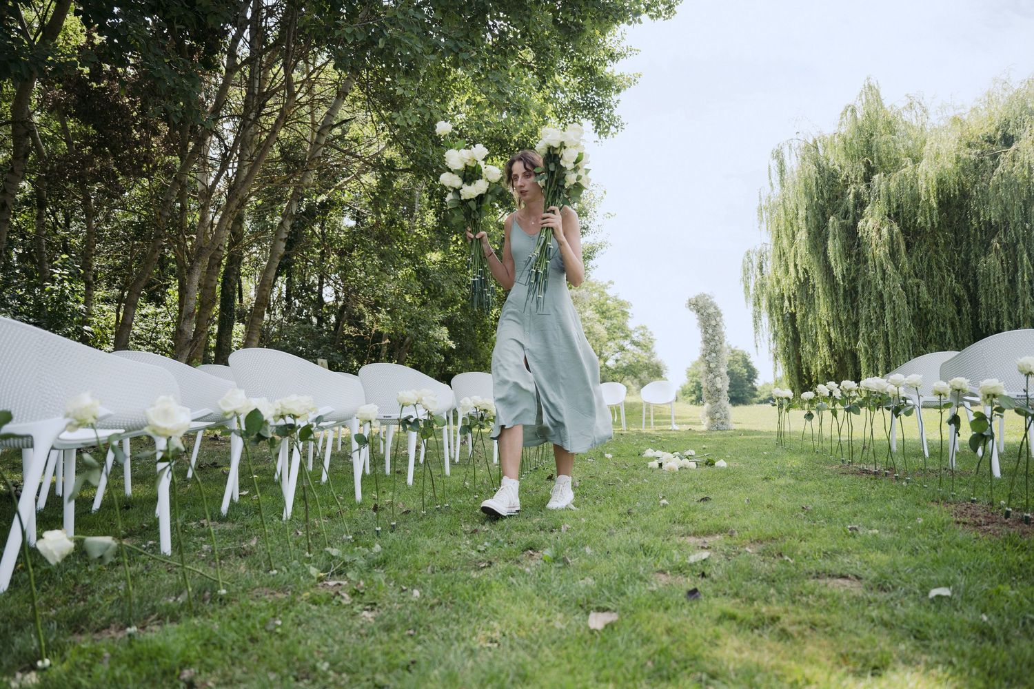 An outdoor wedding ceremony setup with white chairs on a grassy lawn bordered by tall trees and weeping willows.