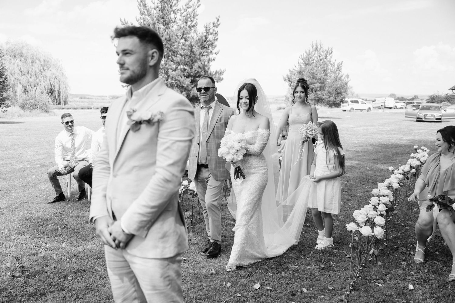 A wedding party in formal attire walks in a procession on a grassy lawn during an outdoor ceremony.