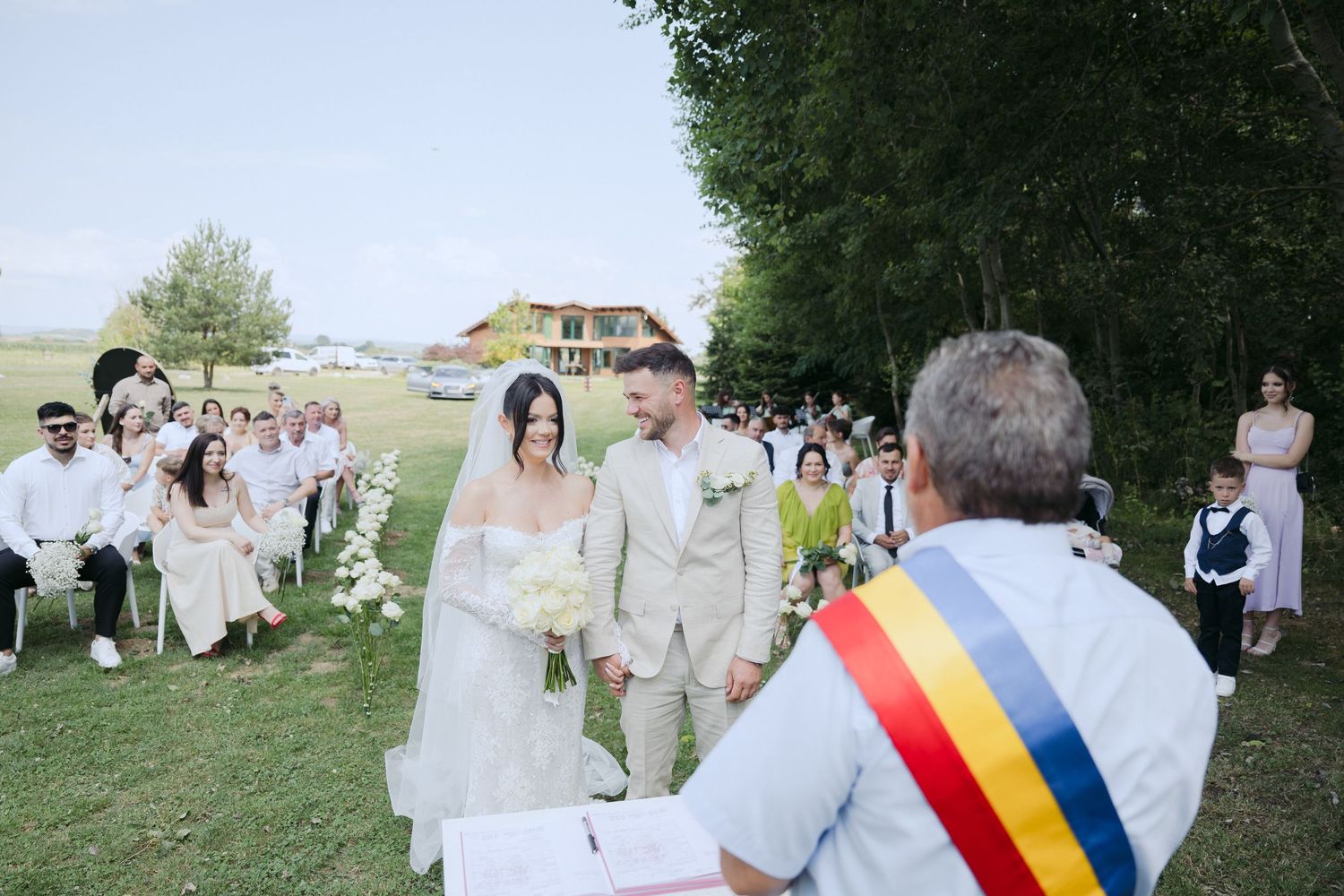 An outdoor wedding ceremony takes place on a grassy hillside with guests seated and an official wearing a tricolor sash.