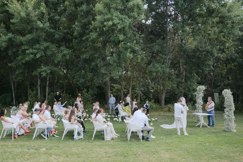 An outdoor garden party with guests seated at white tables amid lush green trees and foliage.