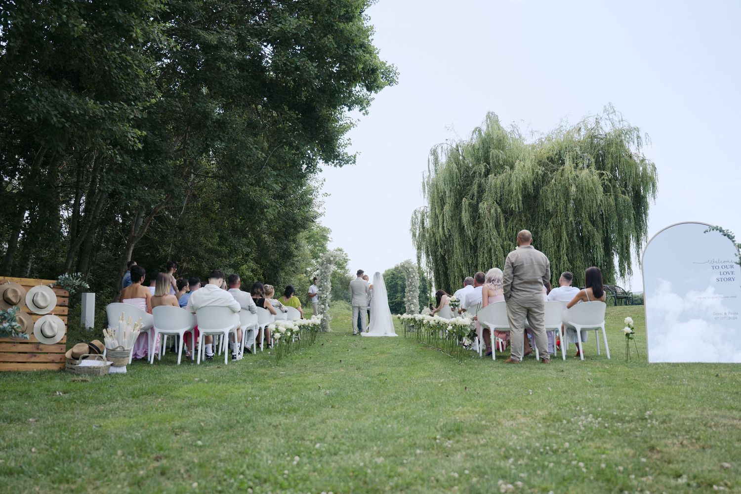 Outdoor wedding ceremony setup on a grassy lawn with white chairs and willow trees in the background.
