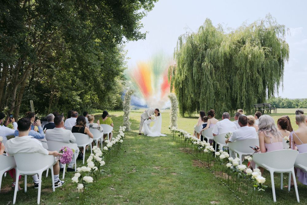 Outdoor wedding ceremony setup with white chairs and colorful smoke bomb creating vibrant rainbow effect in background.