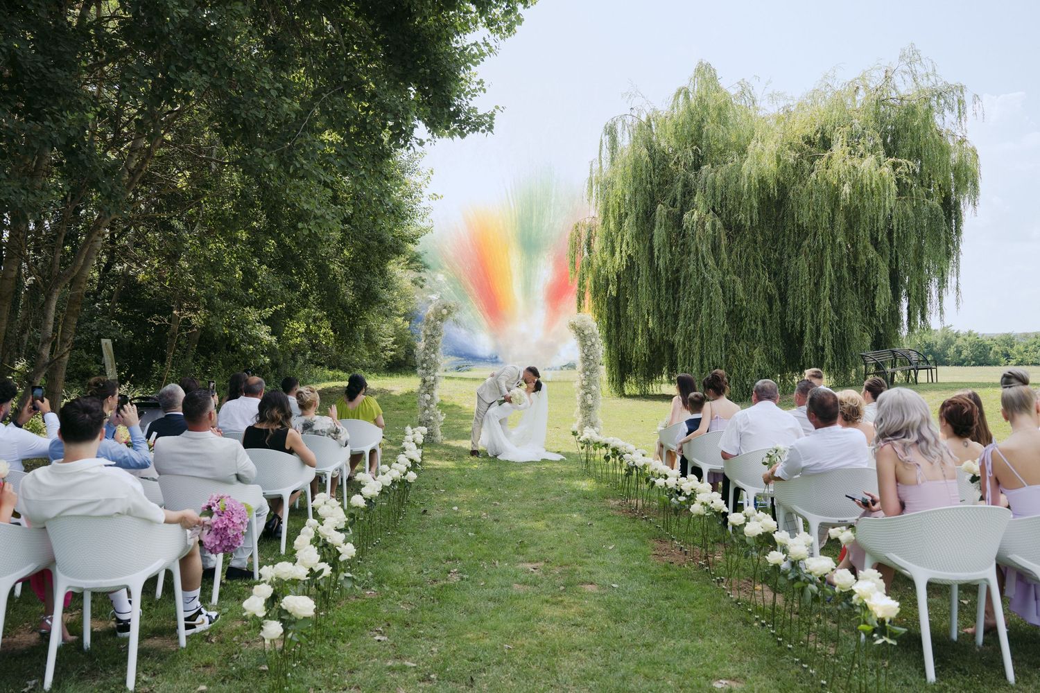 Outdoor wedding ceremony setup with white chairs and colorful smoke bomb creating vibrant rainbow effect in background.
