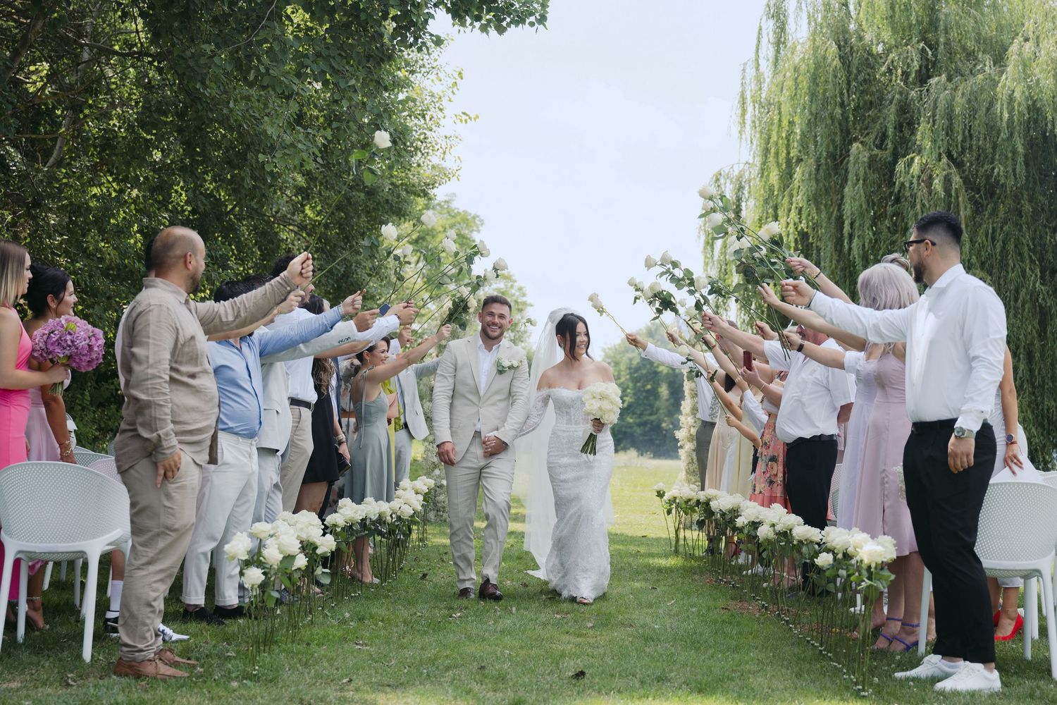 Wedding party attendees create an outdoor walkway arch as two people embrace during a celebration in a lush garden setting.