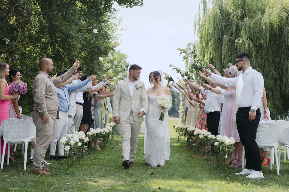 Wedding guests throw rose petals at a couple walking down an outdoor aisle lined with willow trees and white chairs.