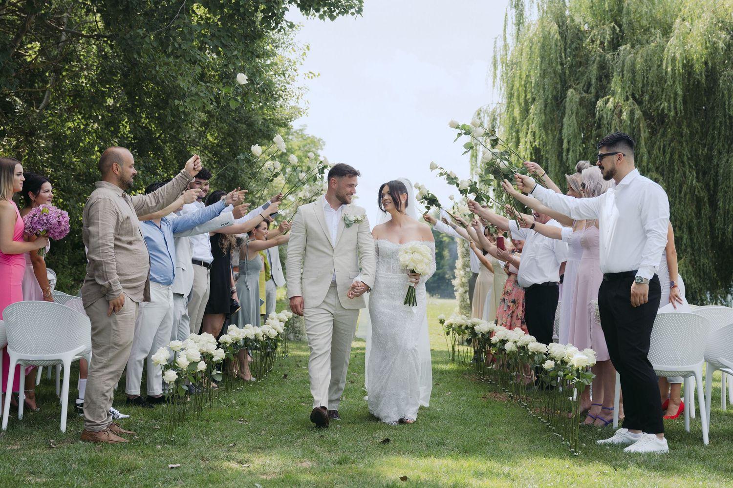 Wedding guests throw rose petals at a couple walking down an outdoor aisle lined with willow trees and white chairs.