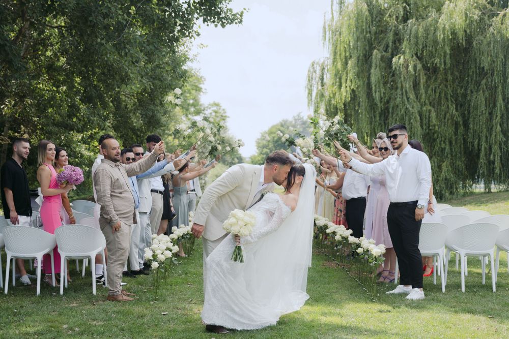 Wedding celebration on a lush lawn with guests lining a path as newlyweds share a romantic dip kiss under willow trees.
