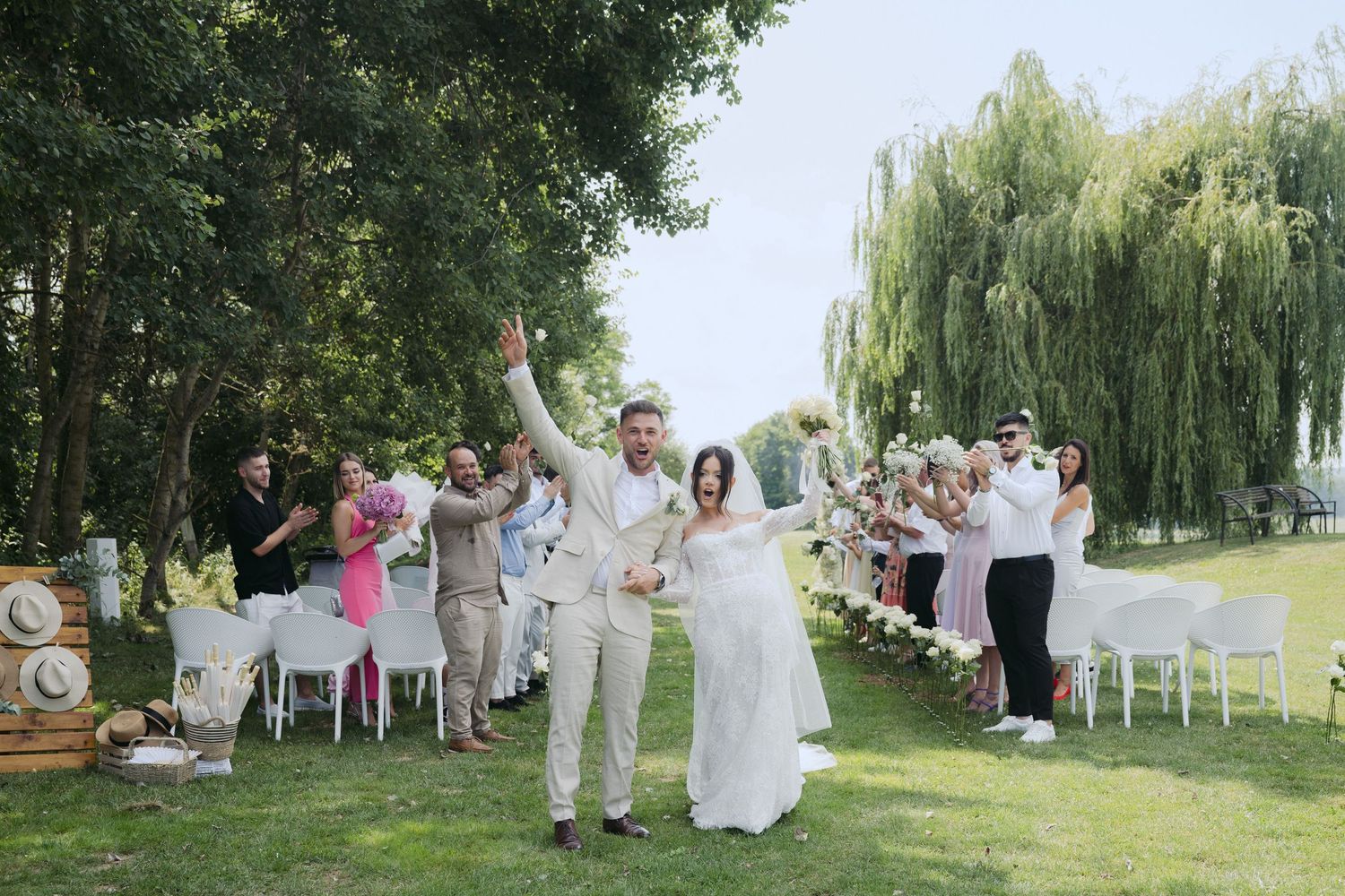 Newlyweds celebrate with guests outdoors as they walk down the aisle lined with white chairs under weeping willows.