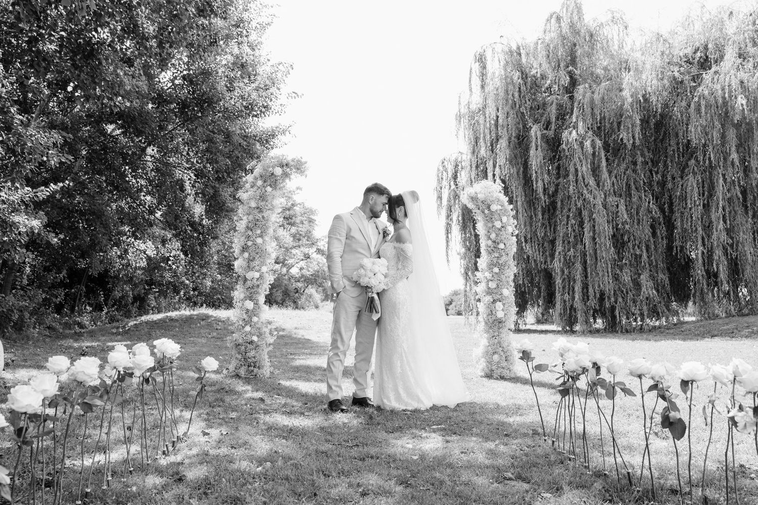 Black and white wedding photo showing a couple embracing beside a lake with weeping willows and garden flowers.