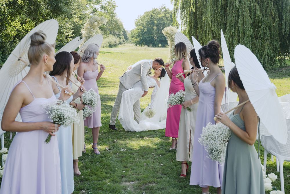 Wedding party gathers outdoors under willow trees on a sunny summer day with bridesmaids holding white bouquets.