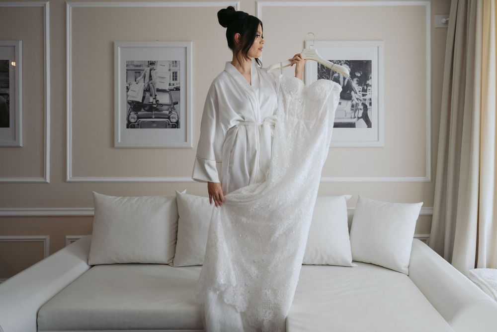 Someone wearing a flowing long white wedding dress stands in an elegant hotel room with framed artwork on the walls.