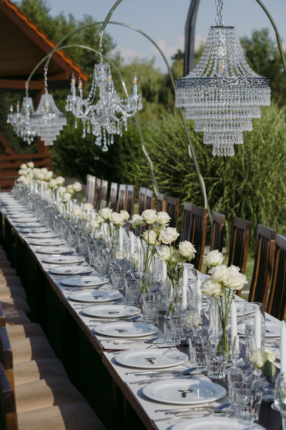 Elegant outdoor wedding table setting with crystal chandeliers and white roses in clear vases along a rustic wooden table.