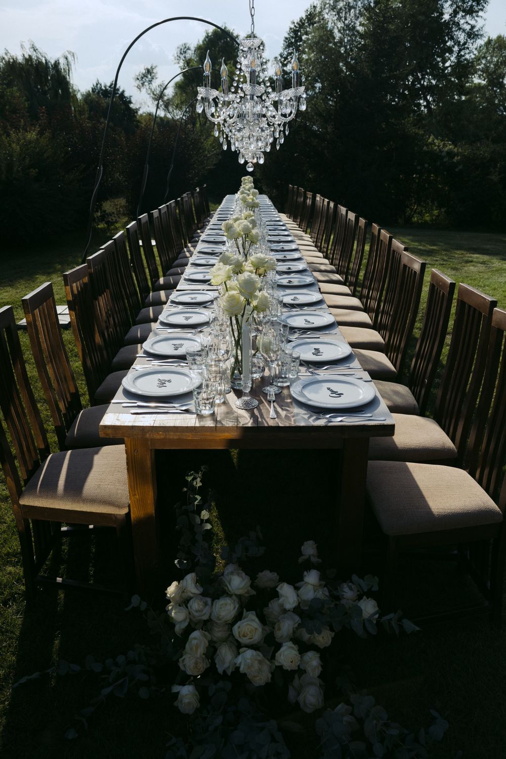 Long rustic dining table with crystal chandelier and white floral centerpieces set for an outdoor garden event.