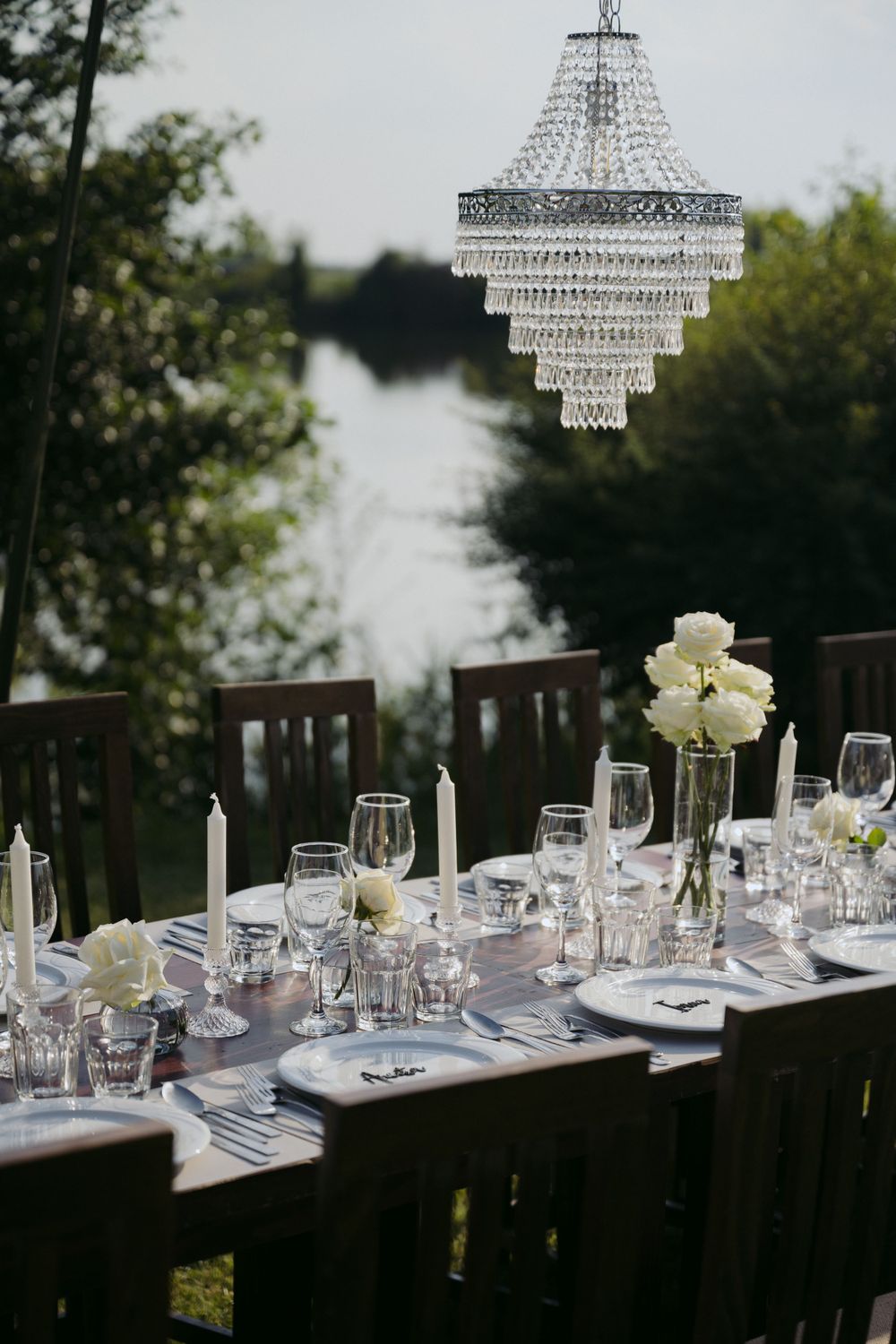 Elegant outdoor table setting with crystal chandelier and white floral centerpiece overlooking lakefront at dusk.