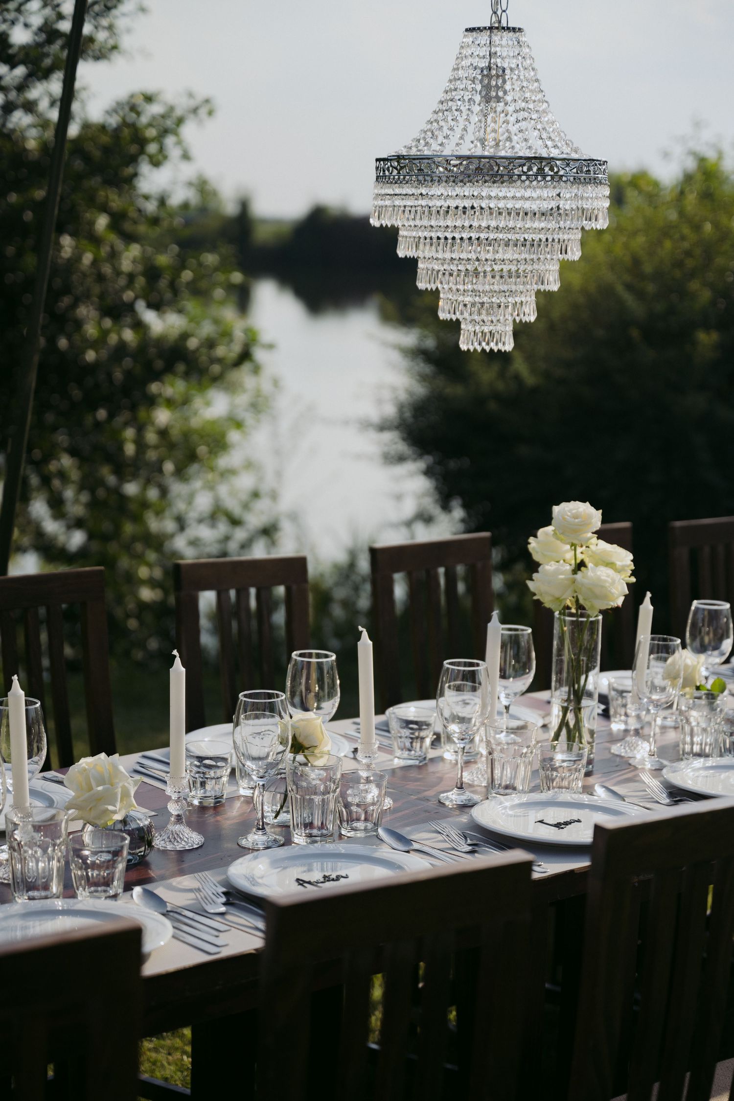 Elegant outdoor table setting with crystal chandelier and white floral centerpiece overlooking lakefront at dusk.