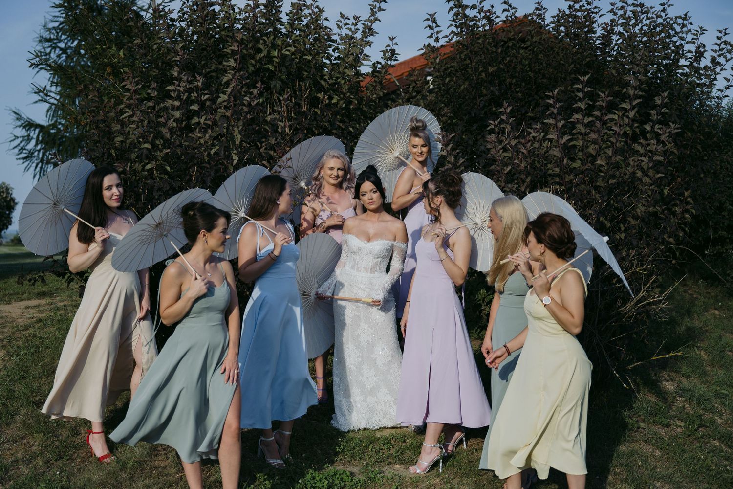 Bridesmaids in pastel dresses holding white umbrellas pose playfully outdoors against greenery.