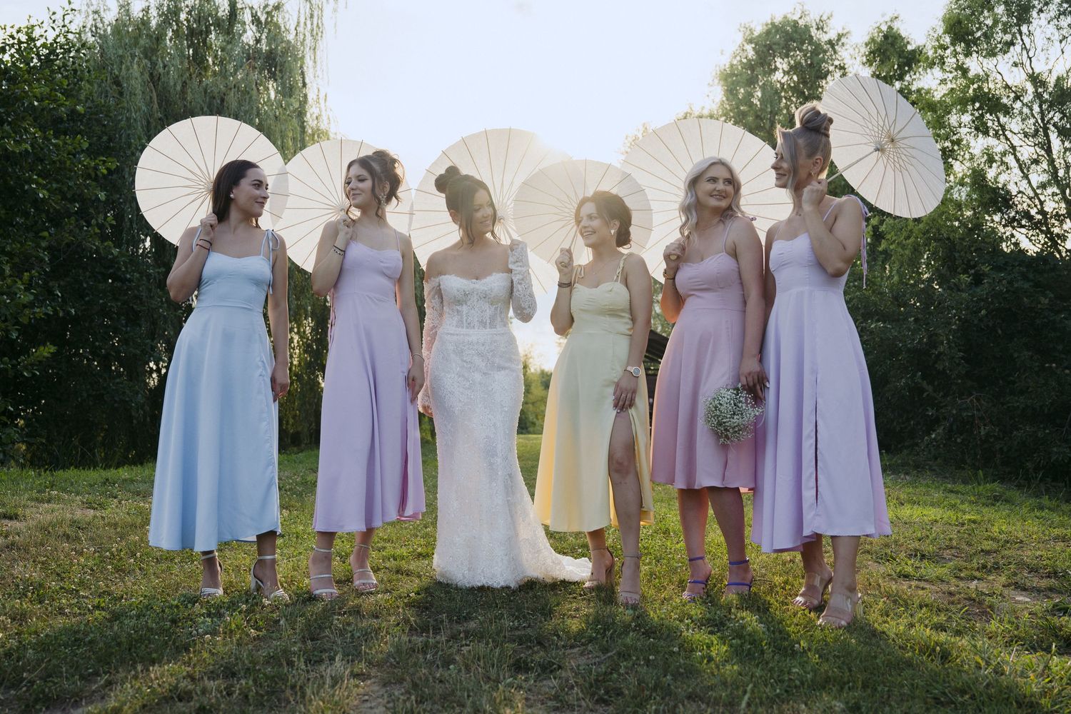 A group stands outdoors on a sunny day holding white parasols and wearing pastel dresses for a wedding party.