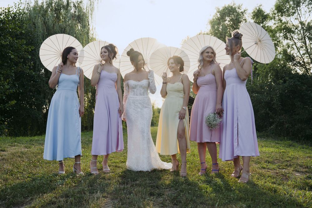 A group stands outdoors on a sunny day holding white parasols and wearing pastel dresses for a wedding party.