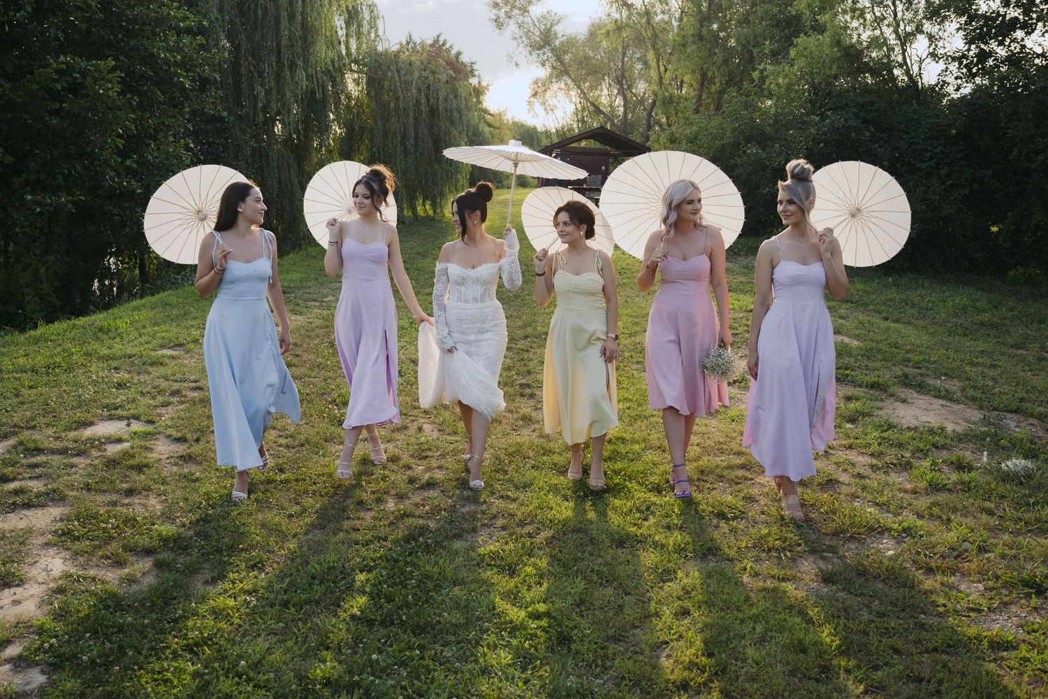 A group in pastel dresses holds white parasols while standing in a sunlit garden during golden hour.