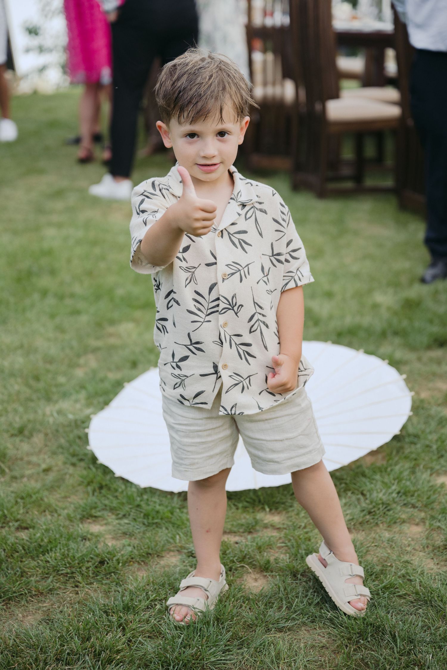 A young child in a leaf-print shirt and khaki shorts stands on grass at an outdoor event, wearing sandals.
