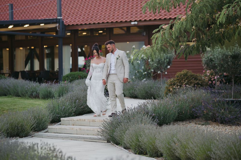 A couple in wedding attire walks together along a stone path bordered by lavender plants at a rustic venue with terra cotta roof.