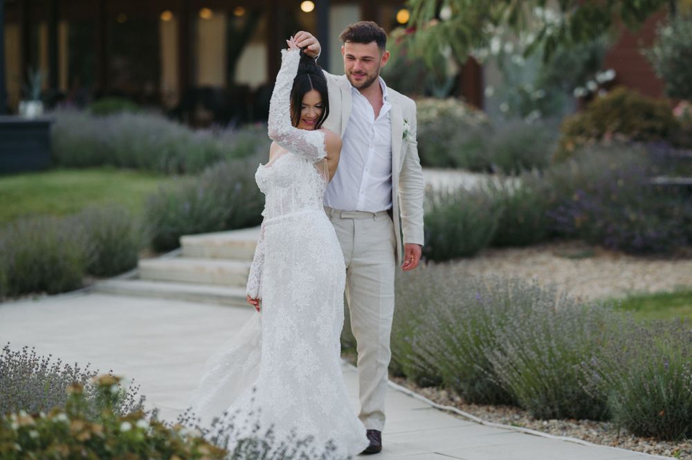 A couple in wedding attire walks down a garden path lined with lavender plants on a summer evening.
