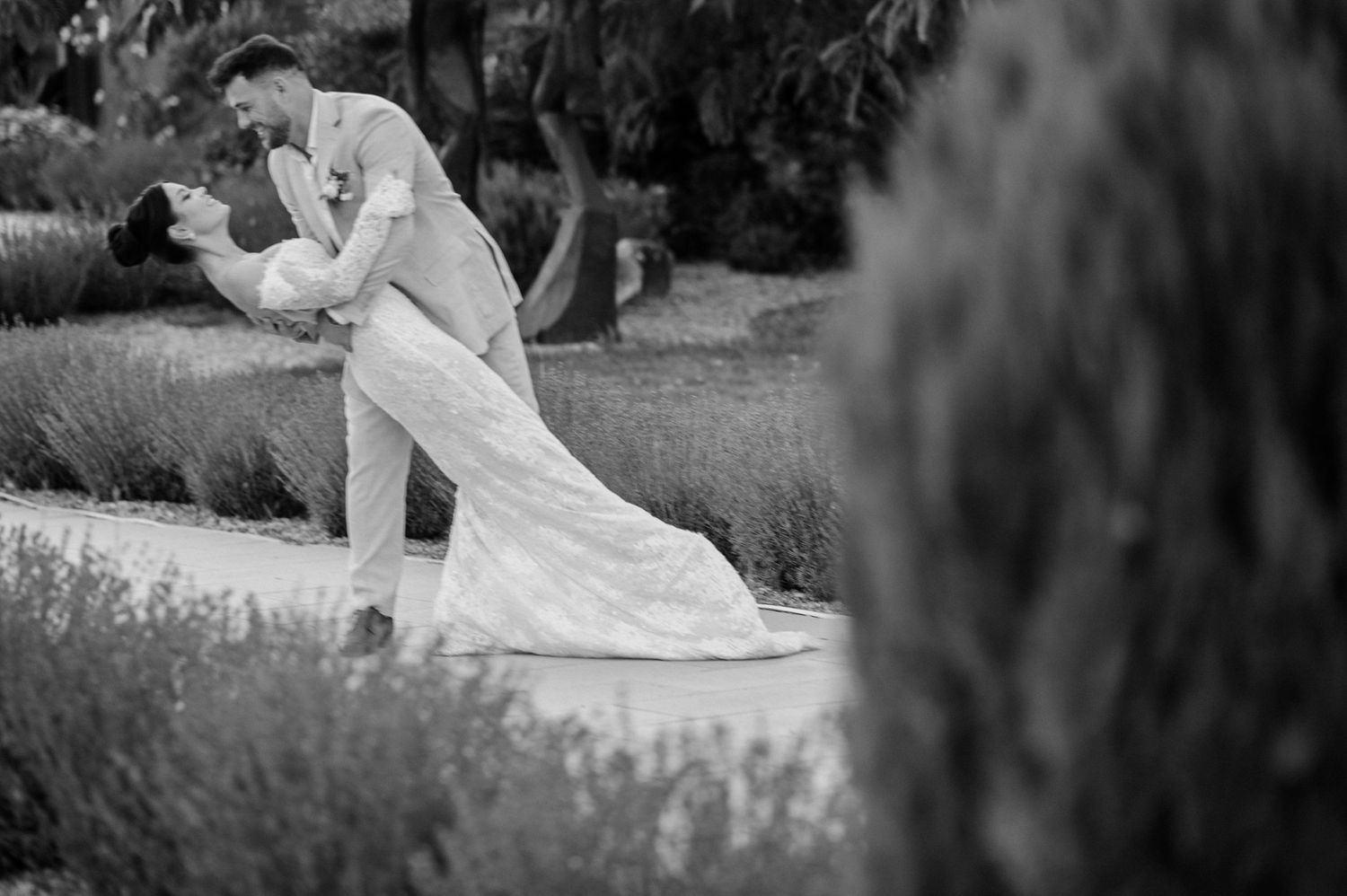 A romantic black and white photograph of a couple in wedding attire embracing in a garden setting.