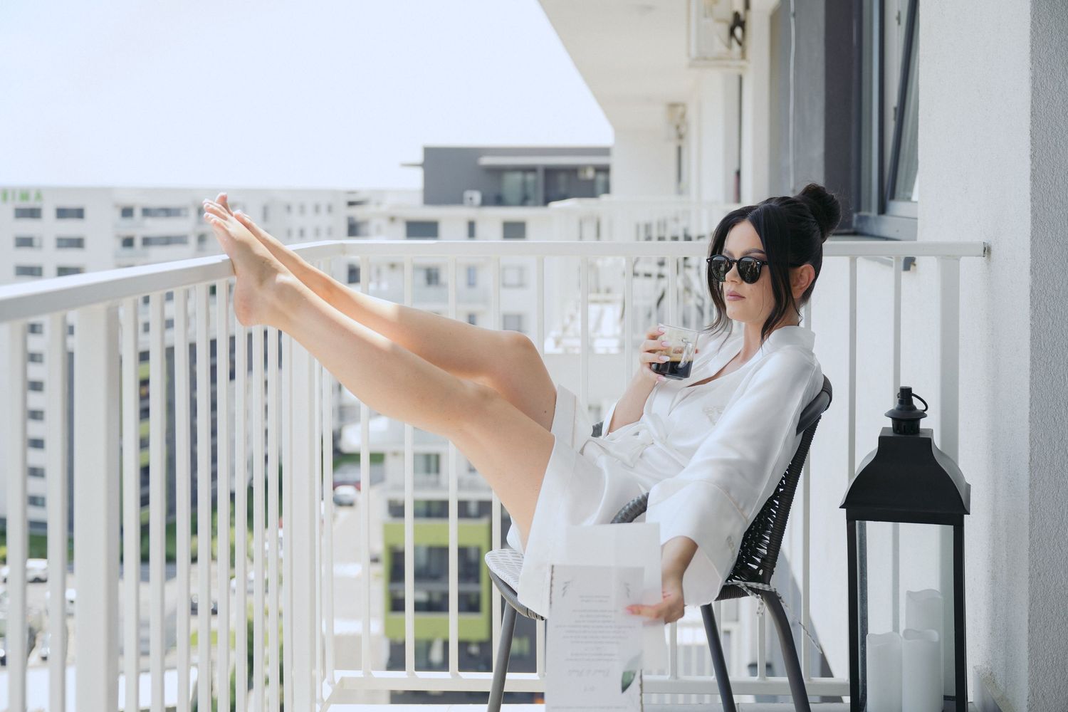 A person in casual white attire relaxes with raised legs on a modern balcony with urban views and a decorative lantern.