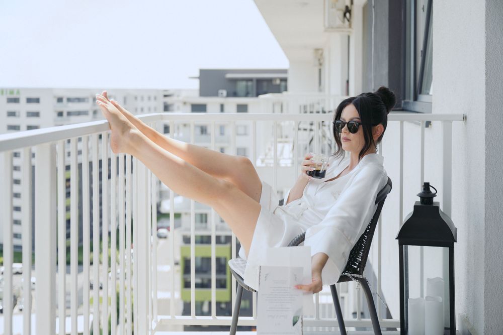 A person in casual white attire relaxes with raised legs on a modern balcony with urban views and a decorative lantern.