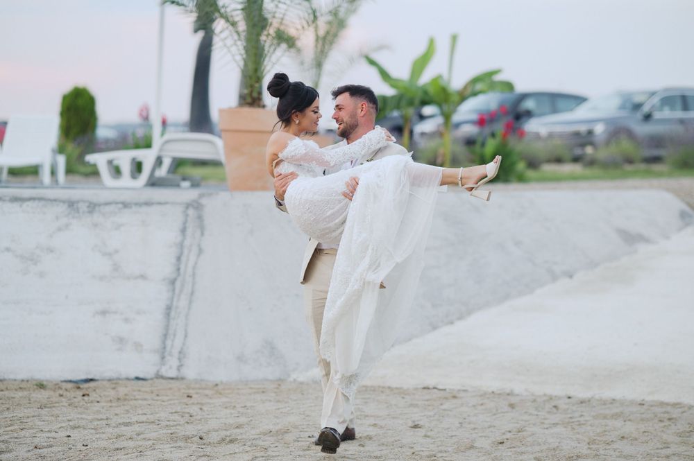 A couple in light colored outfits embraces playfully on a beach near a concrete wall with tropical plants in background.