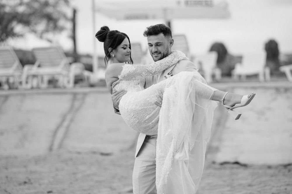A romantic black and white wedding photo shows a couple in a playful moment as he carries her in his arms outdoors.