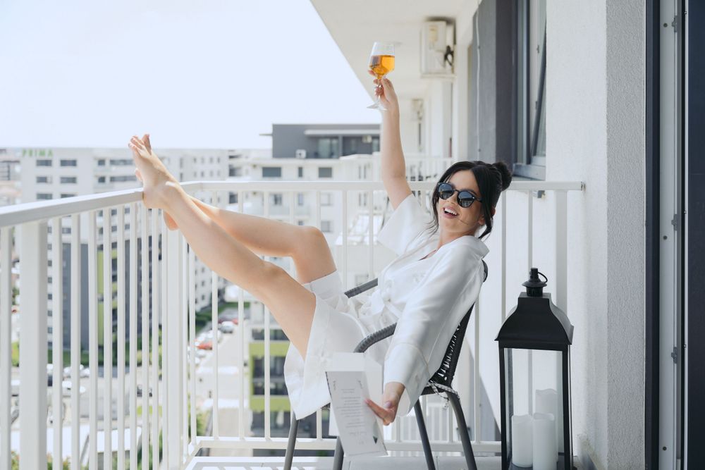 Relaxed person enjoying wine on a balcony with legs up against railing and cityscape view in background.