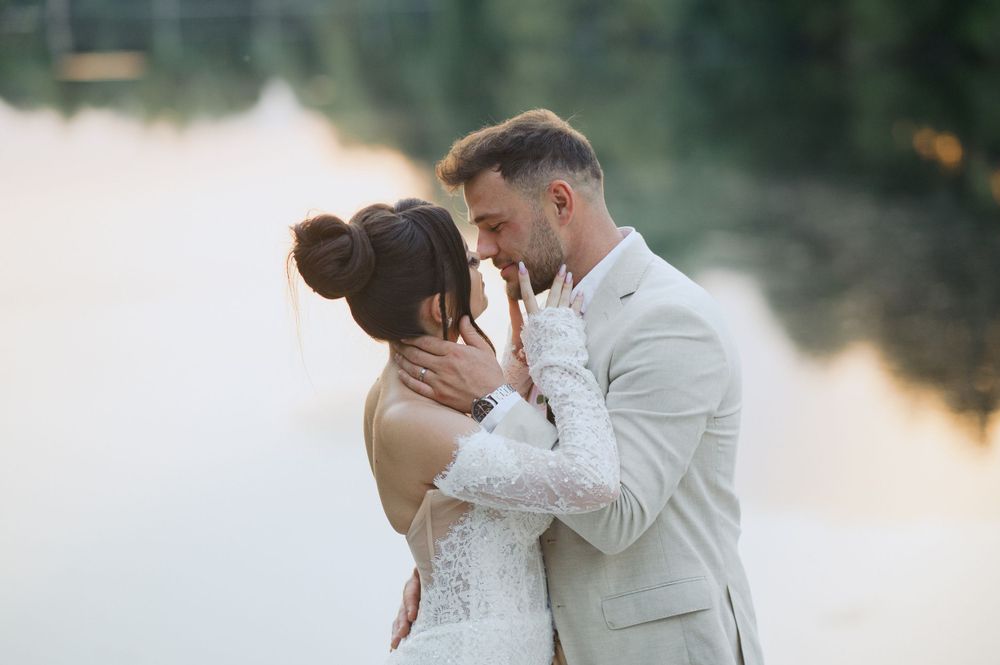 A romantic lakeside wedding photo of newlyweds in white attire embracing during golden hour.