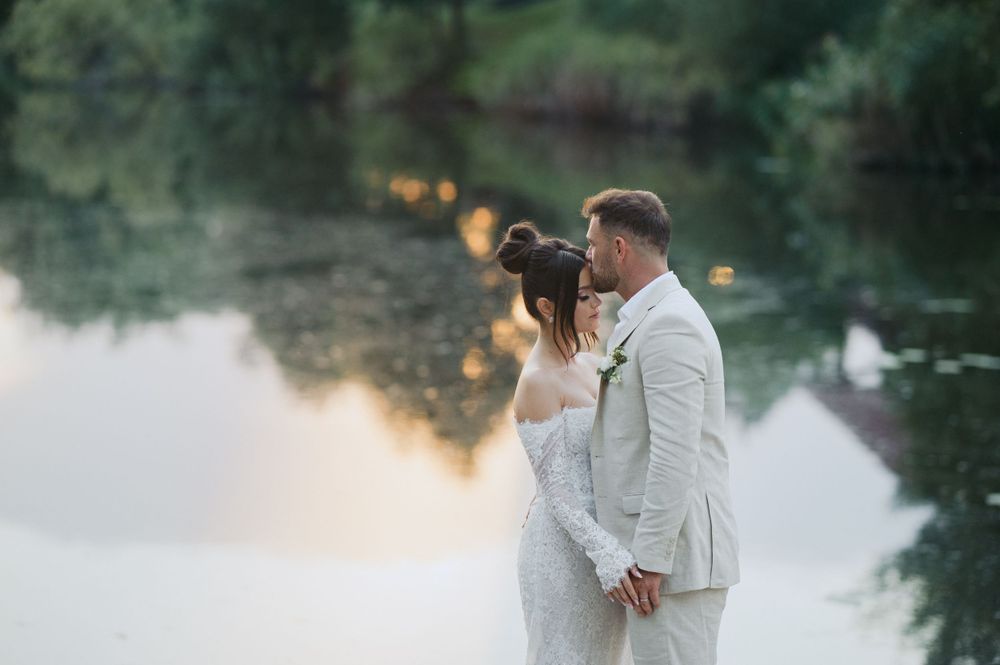 A couple in white attire shares a romantic moment by a misty lake at golden hour.