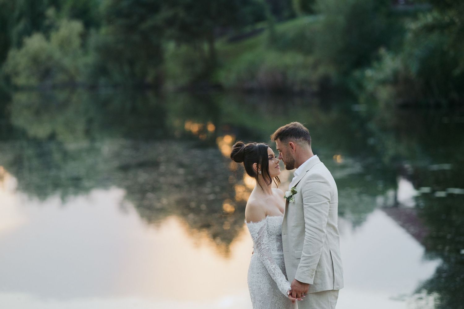 A couple in wedding attire stands close together at sunset by a tranquil lake with golden light reflecting on the water.