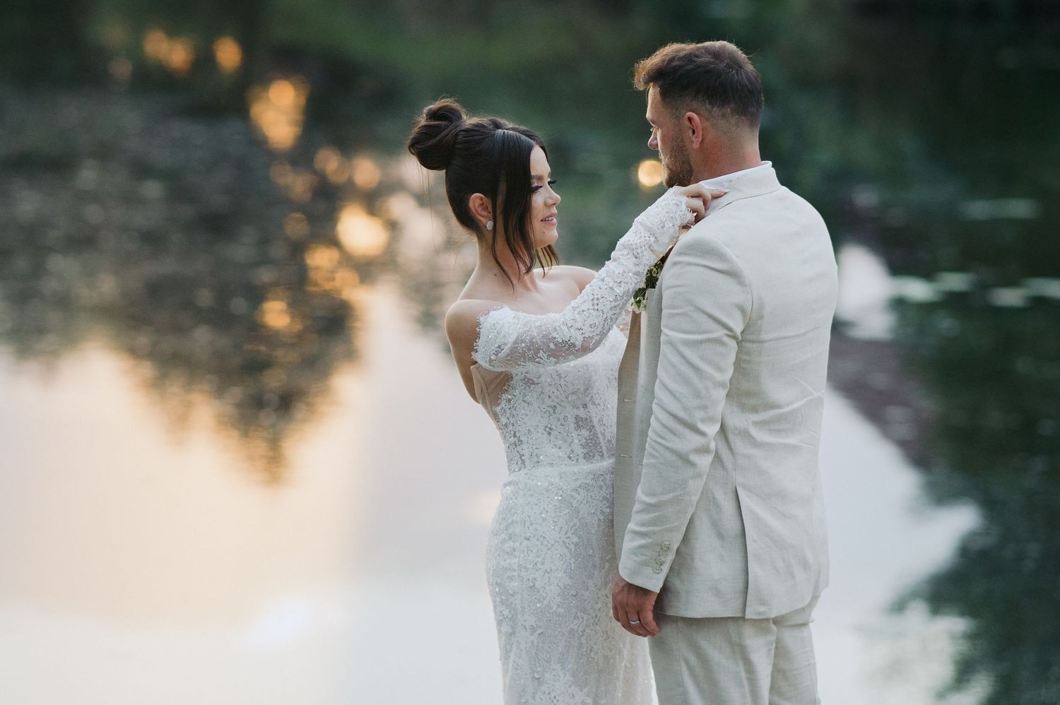 A romantic wedding couple stands by a tranquil lake at sunset with golden light reflecting on the water.
