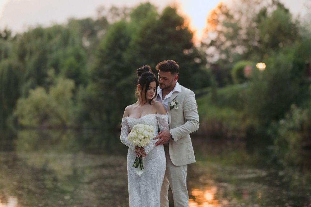 A romantic wedding couple embraces by a lake at sunset while holding a white floral bouquet.
