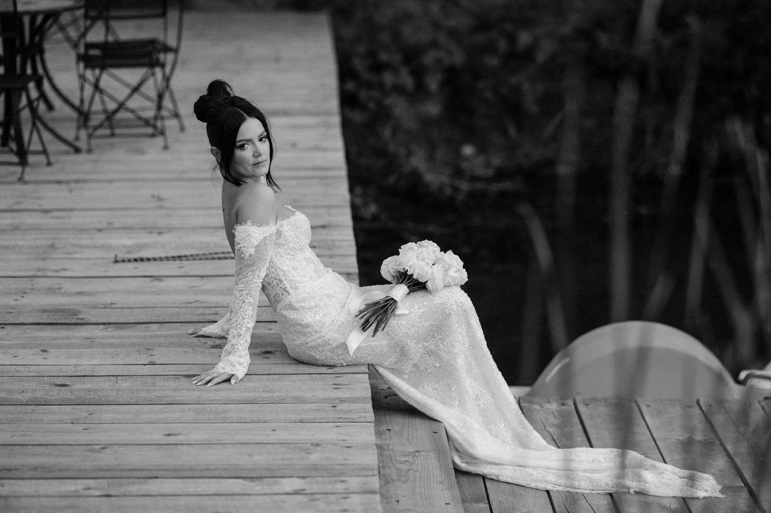 A person in an elegant white dress poses gracefully on a wooden dock by the water.