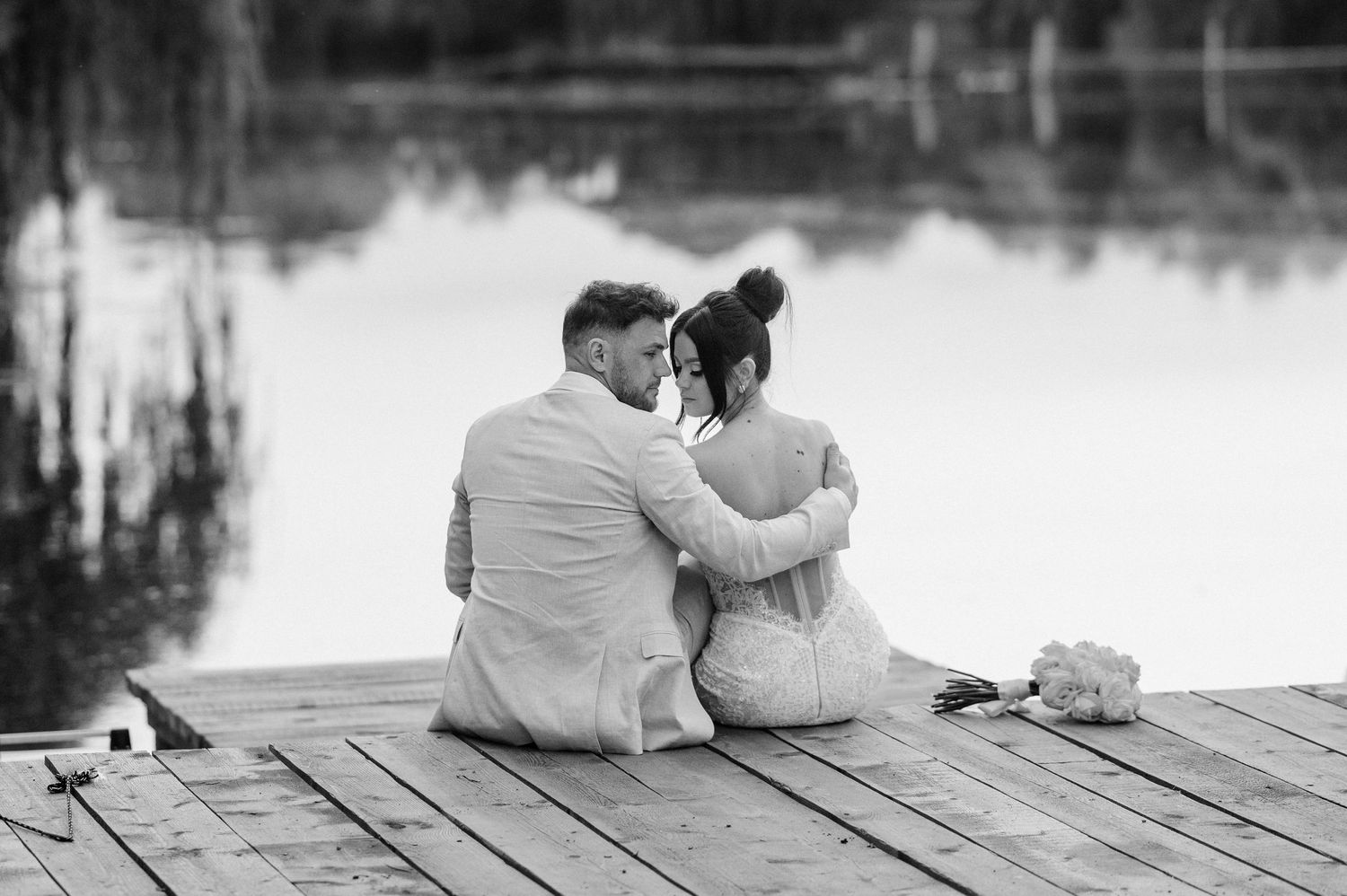 A couple sits intimately on a wooden dock by a tranquil lake in black and white photography.