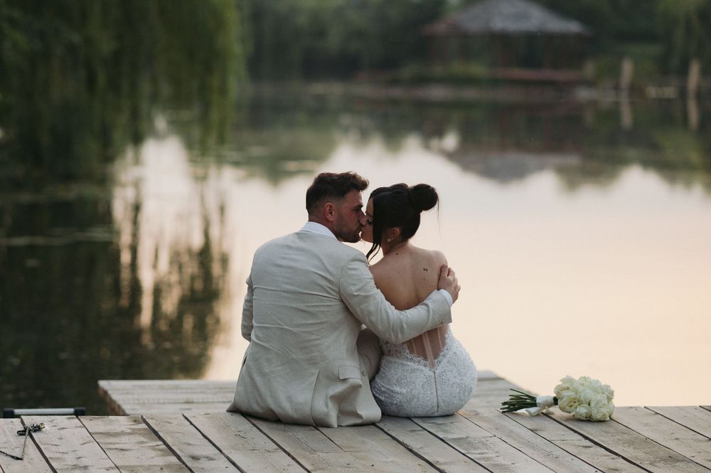 A romantic couple in white sits together on a wooden dock overlooking a serene lake at sunset.