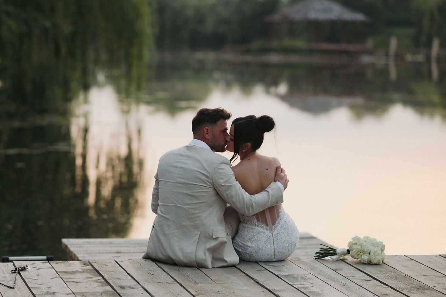 A romantic couple in white sits together on a wooden dock overlooking a serene lake at sunset.