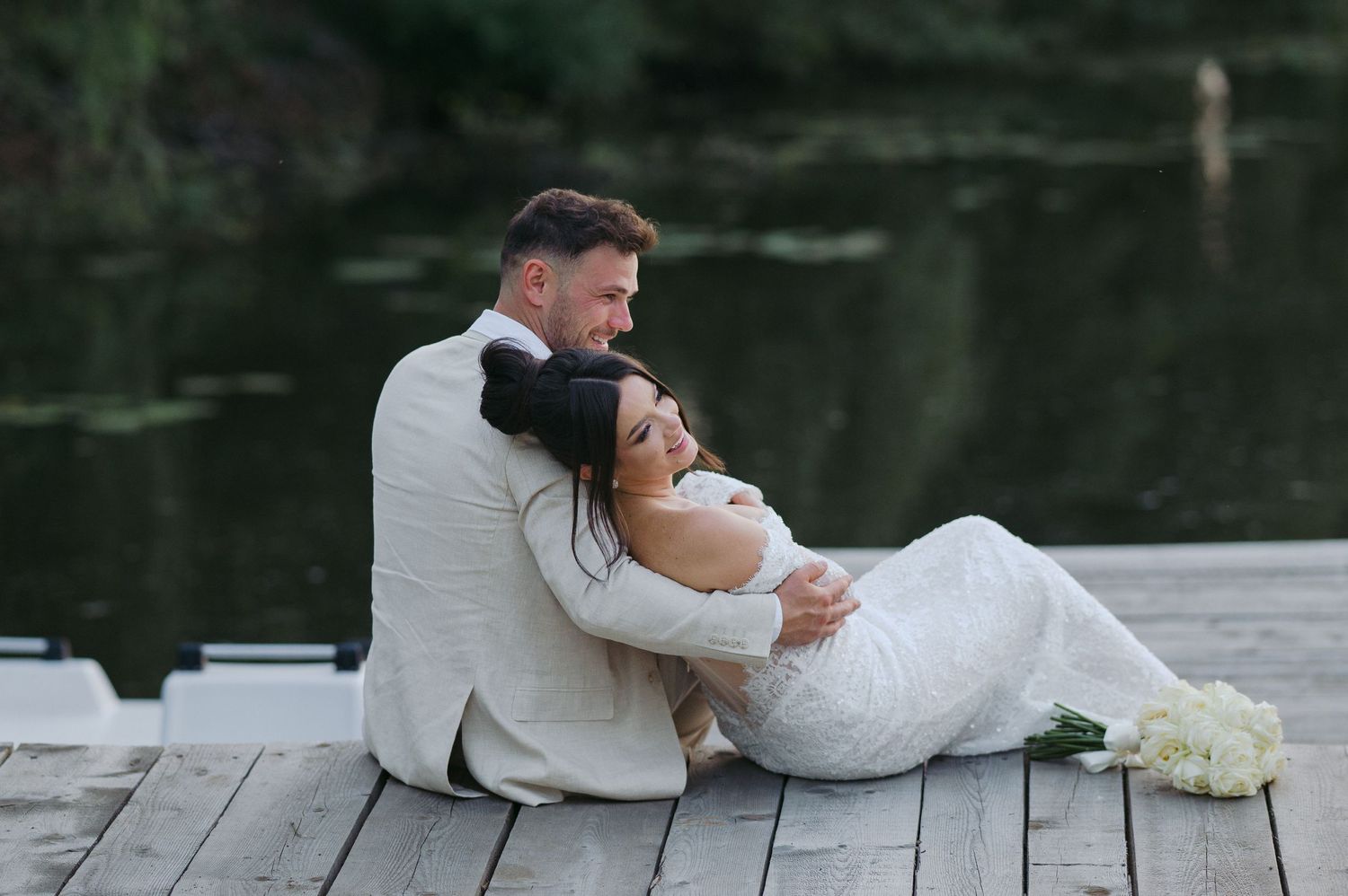 A couple in wedding attire share a romantic moment while sitting on a wooden dock by a tranquil lake.