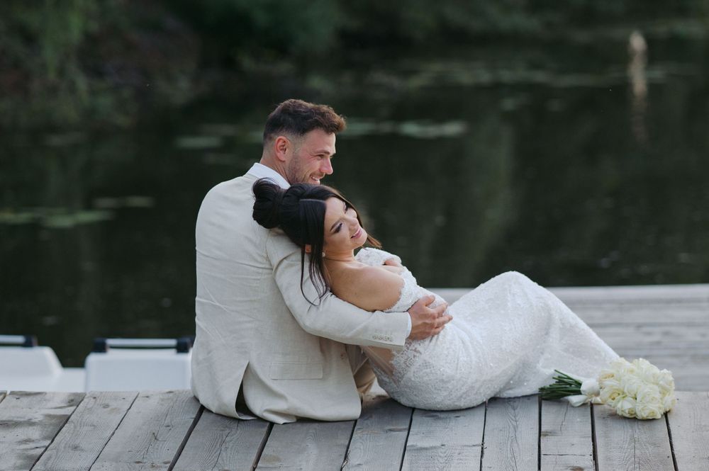 A couple in wedding attire share a romantic moment while sitting on a wooden dock by a tranquil lake.